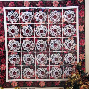 May include: A quilted Advent calendar featuring 25 white crocheted doilies with red poinsettia flowers in the center. The doilies are arranged in a grid pattern on a red and green poinsettia fabric background. The text "Quilted Advent Calendars" is at the top of the image. The text "Designs by Sandra L. Hatch" is below the calendar. The text "Poinsettia Bouquet" is below the calendar. The text "5 quick & easy Advent calendars help you celebrate Christmas" is at the bottom of the image.