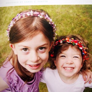 May include: Two girls wearing colorful headbands made with beads and buttons. The headband on the girl in the front is made with pink, white, and purple beads. The headband on the girl in the back is made with red, green, and yellow buttons.