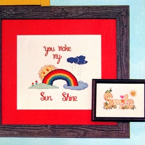 May include: Two framed cross-stitch embroidery designs with a sunny theme. The first design says "Have a Sunshiny Day" with a yellow sun and a blue cloud. The second design says "You make my Sun Shine" with a rainbow, sun, and clouds.