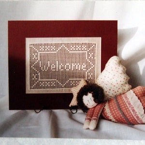 Peut inclure: Un motif de point de croix encadré avec les mots "Welcome Home" dans un cadre blanc avec un passe-partout rouge. Une petite poupée avec une robe rose et blanche est couchée sur une surface blanche.