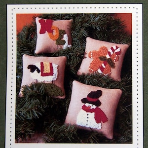 May include: Four needlepoint Christmas pillow ornaments, each measuring 4 1/2 inches square. The ornaments feature a snowman, a gingerbread person, a lamb, and the word "Noel". The ornaments are displayed on a bed of green pine branches.
