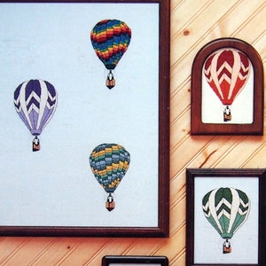 May include: A collection of six framed cross-stitch artworks featuring hot air balloons, labelled Silver Lining Creations and Cloud Dancers 1. The designs include various colourful balloon patterns displayed on a wall above a picnic basket and food.