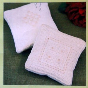 May include: A package for a box pincushion kit with the text "Silkstone Design". The package features two white, square, crocheted pincushions. One has a floral design, and the other has a geometric pattern. The background is green.