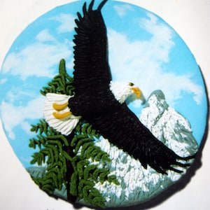 May include: A ceramic wall plaque featuring a bald eagle in flight against a blue sky with white clouds. The eagle is perched on a green tree branch with snow-capped mountains in the background.