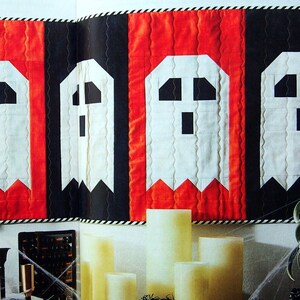 May include: A Halloween quilt with five white ghost figures on a black and red background. The quilt is hanging on a wall with candles and a plant in front of it.