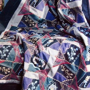 Peut inclure: Une courtepointe en patchwork avec une bordure noire, présentant une variété de motifs de broderie florale et de ruban. La courtepointe est pliée et drapée sur un banc en bois.