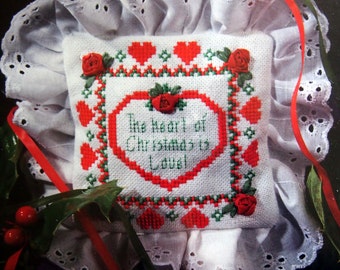 Könnte beinhalten: Ein weißes Stoffkissen mit einem Kreuzstich-Design aus rotem Herzen und grünen Blättern. Das Kissen hat ein rotes Band und eine weiße Spitzenborte. Der Text auf dem Kissen lautet "The heart of Christmas is Love!"