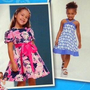 May include: Simplicity 2989 sewing pattern for girls' dresses. The pattern features two dress designs, one with a floral print and pink sash, and another with a blue and white polka dot pattern. The package is inspired by Project Runway.