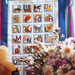 May include: A white fabric alphabet chart with colorful embroidered animals and letters. Two teddy bears, one holding a teacup, are sitting in front of the chart with a bouquet of flowers.