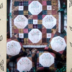 Puede incluir: Un patrón de colcha para una colcha y almohadas en forma de cesta. La colcha está hecha de tela de retazos en tonos de marrón, rojo, verde y blanco. Las almohadas también están hechas de tela de retazos y presentan cestas de flores bordadas.