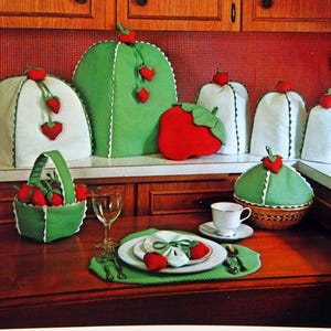 May include: A collection of fabric kitchen accessories featuring strawberry motifs, including covers for appliances, a basket, a placemat, and napkins. The text Strawberry Kitchen Kollection and The Gingham Goose Patterns & Pretties, Inc. is visible.