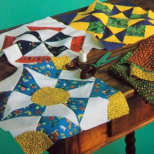 Op de afbeelding: De omslag van een boek met de titel "State Capitals Quilt Blocks" wordt weergegeven, samen met verschillende kleurrijke patchwork quiltblokken. De boekomslag is groen met witte tekst. De quiltblokken liggen op een houten tafel. De tekst op de omslag luidt "50 Patchwork Patterns from Hearth & Home Magazine."