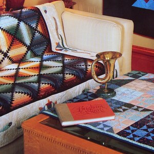 May include: A living room interior with a couch covered in a colorful quilt. A coffee table with a book titled "Brigham Street" and a quilt top on it. A sign on the wall reads "Vanessa-Ann's Living with Quilts".