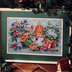 May include: A cross-stitch pattern featuring a framed image of a blue and white birdhouse with a red roof, surrounded by flowers and a nest with eggs. The pattern is titled "Summer Homes" and is by Barbara Mock.