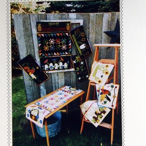 May include: A set of quilted wall hangings and table runners with a variety of seasonal themes. The hangings feature designs like a welcome sign, a bird, and a sunflower. The table runners feature designs like a teapot, a bird, and a sunflower. The hangings and runners are all made in a patchwork style with a variety of fabrics and colors. The text "Snuggles Quilts by Deanne Eisenman™" is visible at the bottom of the image.
