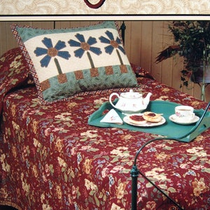 Op de afbeelding: Een quiltpatroonboek getiteld "Fancy Free" van Thimbleberries, met een rode bloemenbedsprei met een groene dienblad en een theeservies erop. De omslag van het boek toont ook een kussen met een bloemenontwerp.