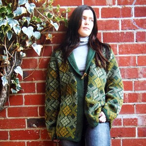 May include: A woman wearing a green and yellow patterned cardigan with a high collar and a white turtleneck underneath. She is wearing blue jeans and black boots. The woman is standing in front of a brick wall.