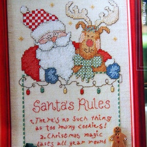 Puede incluir: Obra de arte navideña bordada a punto de cruz en un marco rojo. Papá Noel y un reno están representados encima del texto "Santa's Rules". Las reglas están bordadas con hilo rojo sobre un fondo color crema. Un hombre de jengibre y un regalo están adjuntos a la obra.