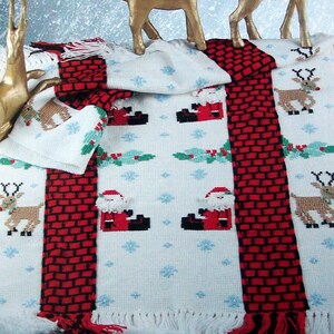 May include: White knitted throw blanket with red and black stripes and a Christmas theme. The blanket features a repeating pattern of Santa Claus, reindeer, and snowflakes. Two gold reindeer figurines are placed on top of the blanket.