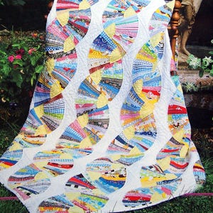 May include: A vibrant quilt with a fan design, displayed in a garden. The quilt has a white base with colourful, segmented fabrics. The words "Heirloom Quilts" are visible at the top. The scene is framed by a pink border.