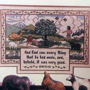 May include: Framed cross-stitch artwork depicting a biblical scene with text from Genesis. The artwork features a tree, animals, and a figure, with a decorative border. Miniature animal figurines are in the foreground.