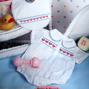 Puede incluir: Un pelele blanco para bebé con un estampado de corazones azules y rosas y un babero a juego. El pelele tiene un cuello Peter Pan y mangas cortas. El babero tiene un borde festoneado y un estampado de corazones azules y rosas. El conjunto se muestra en una cesta de mimbre blanca con una manta azul y blanca.