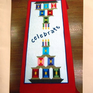 May include: A white quilted table runner with a red border. The runner features a design of stacked birthday cakes with candles and the word "celebrate" in blue lettering. The runner is on a wooden table. "Original Design By K & H Quilt Shoppe" is printed at the bottom of the image.
