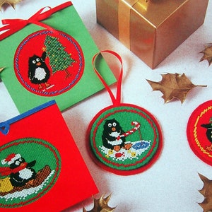 May include: Four handmade Christmas decorations featuring penguins in different festive scenes. The decorations are round and have a green background with red trim. The penguins are wearing hats, scarves, and holding gifts.