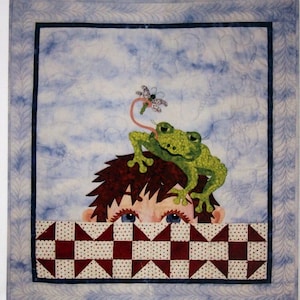 May include: A fabric quilt with the text "Frog Days of Summer" and a whimsical design. A green frog with a long tongue sits atop a child's head, reaching for a dragonfly. The quilt measures 30" x 32".