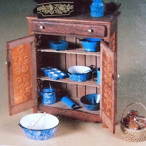 Puede incluir: Un armario de madera con un solo cajón y dos puertas. El armario está lleno de vajilla de esmalte azul y blanco, incluyendo cuencos, ollas y un rallador. El armario tiene una puerta decorativa con un diseño de corazón. Hay una pequeña caja de madera con un diseño floral en la parte superior del armario.