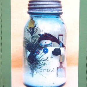 May include: A clear glass jar with a blue metal lid, containing a snowman figurine, a small shovel, and a sprig of evergreen. The jar has the words "Let it Snow" written on a white sign. The words "Threads That Bind" are at the top.