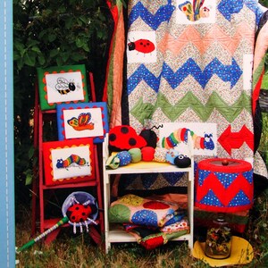 May include: A display of handmade items with a quilt, framed insect embroidery, and plush toys. The quilt features a chevron pattern with ladybug and butterfly appliqués. The framed embroidery includes a bee, butterfly, and caterpillar. The text "ADORNit" is visible.