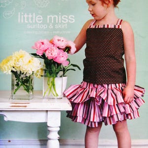May include: A sewing pattern titled little miss suntop & skirt by carina gardner featuring a brown polka dot tank top and a multi-tiered ruffled skirt with vertical stripes in shades of pink, red, black, and white on a light blue background.