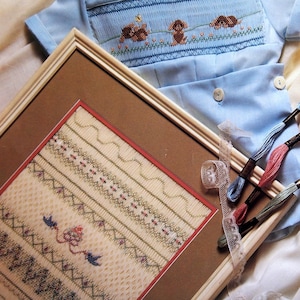 May include: A light blue baby outfit with a smocked bodice featuring embroidered dachshunds. A framed needlepoint sampler with various stitch patterns and a selection of embroidery floss are also present. The image also includes the text "ellen mccarn".