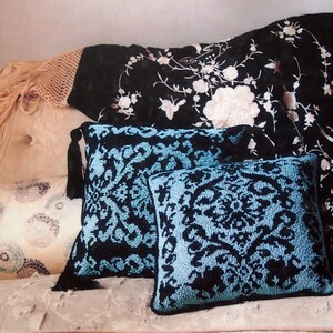 May include: A book titled "glamour knits at home" by Erika Knight is displayed on a floral patterned sofa. Two square pillows with a black and blue floral design are in front of the book. The pillows have black tassels. A black floral patterned throw is behind the pillows.