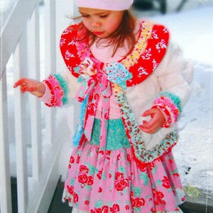 May include: A young child wearing a white jacket with a red floral collar and cuffs, a pink dress with a floral pattern, and a matching pink hat. The outfit is part of the "Butterfly Kisses" patterns collection.