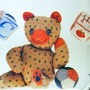 Puede incluir: Un oso de peluche marrón con nariz roja y ojos azules se sienta sobre un fondo blanco. El oso sostiene una pelota multicolor en su pata derecha. Hay tres bloques bordados en el fondo, uno con una flor y la letra 'B', uno con una manzana roja y la letra 'A', y uno con una letra azul 'C'. La imagen es de un patrón de costura vintage para un proyecto artesanal. El número del patrón es 4685.