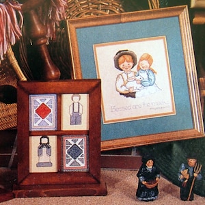 May include: Three wooden frames with cross-stitch embroidery. The centre frame features a cross-stitch design of two children holding a white rabbit with the text "Blessed are the peacemakers". The other two frames feature geometric patterns.