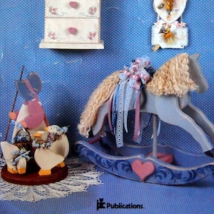 May include: A blue rocking horse with a white mane and tail, a pink bow, and a pink heart on the base. There is a white dresser with pink hearts and flowers on the drawers, and a wall hanging with three paper hearts featuring vintage illustrations of children.