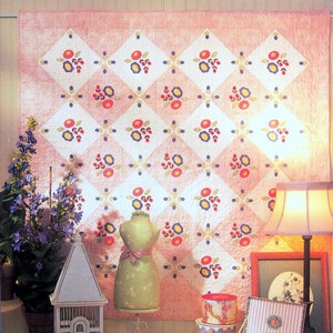 May include: A pink and white quilt with a floral pattern, titled "Blossom Hill". The quilt is displayed on a wall in a room with a white birdcage, a mannequin, and various decorative boxes.
