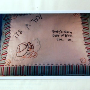 May include: A white pillow with a red, white, and brown striped border. The pillow is embroidered with the text "It's a Boy" and a small baby wearing a hat. The pillow also has a space for the baby's name, date of birth, and weight.