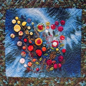 Op de afbeelding: Een blauwe, gestikte wandhanger van stof met een bloemenmotief. De bloemen zijn gemaakt van verschillende gekleurde stoffen cirkels en zijn in het midden van het stuk in een cluster gerangschikt. De achtergrond is donkerblauw met een lichtblauw swirl-patroon.