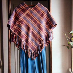 May include: A plaid poncho in shades of purple, orange, and brown with fringe, hanging above blue pants. The poncho is part of a weaving project. The text on the image says "Autumn Sunset Poncho by Inci Koker."
