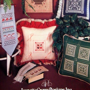 Könnte beinhalten: Eine Sammlung von Kreuzstich-Stickmustern mit verschiedenen Motiven, darunter eine Schale mit Früchten, ein Willkommensschild und ein Blumenmuster. Die Muster sind auf Kissen, einem Lesezeichen und einer Wandbehänge dargestellt. Der Text auf dem Willkommensschild lautet "Willkommen in unserem Zuhause". Jeanette Crews Designs, Inc. 4 Killian Hill Rd. Lilburn, Ga. 30247