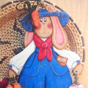 May include: Wooden bunny figure wearing a blue dress with white sleeves and a red scarf. The bunny has pink ears and cheeks, and holds carrots and a bundle of flowers. The figure is decorated with a blue hat and a tag that says "CARROT SEEDS".