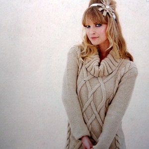 May include: A beige knitted sweater with a cable knit pattern and a cowl neck. The sweater is worn by a model with blonde hair and a headband. The text "The second Sublime Aran hand knit book" is printed on the bottom of the image.