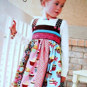 May include: A young girl wearing a colorful cupcake-themed dress. The dress features a white long-sleeved top, brown polka-dot straps, and a patchwork skirt with cupcake and floral patterns. The text "the CUPPYCAKE dress" is visible.