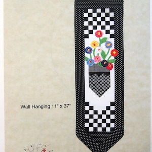 May include: A wall hanging featuring a pocket design filled with colorful posies. The design includes a black and white checkered pattern, a black and white polka dot pattern, and the text "POCKET FULL OF POSIES". The wall hanging measures 11" x 37".