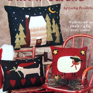 May include: A book titled "Pillow Parade" by Cathy Pendleton, featuring a collection of 16 handmade pillows for each season. The book cover shows a variety of pillows with different designs, including a house, a dog, a sheep, and an angel. The pillows are displayed on wooden chairs and a rocking chair.