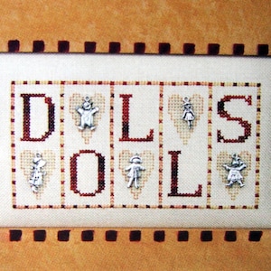 Peut inclure: Broderie au point de croix avec le mot "DOLLS" en lettres rouges et brunes, entourée de cœurs avec de petits charmes de poupées. La broderie est encadrée dans un cadre brun et blanc avec une bordure brune.